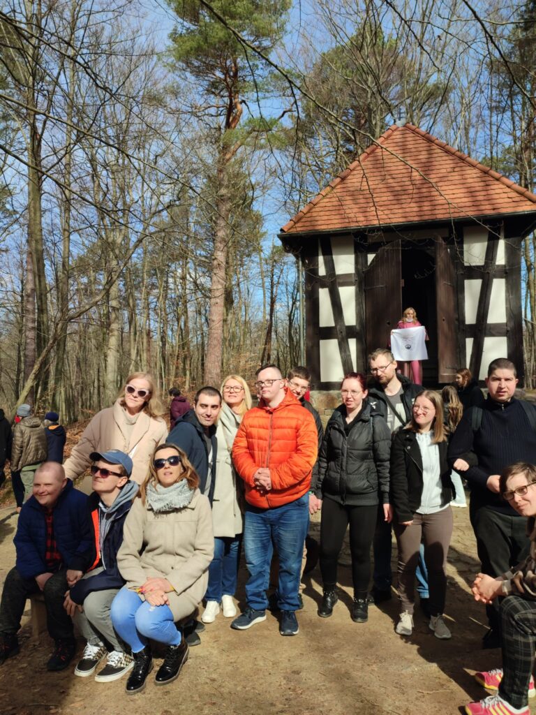 Grupa uczestników Środowiskowego Domu Samopomocy w Wejherowie stojąca przed kapliczką w lesie w słoneczny dzień, podczas obchodów Misterium Męki Pańskiej.