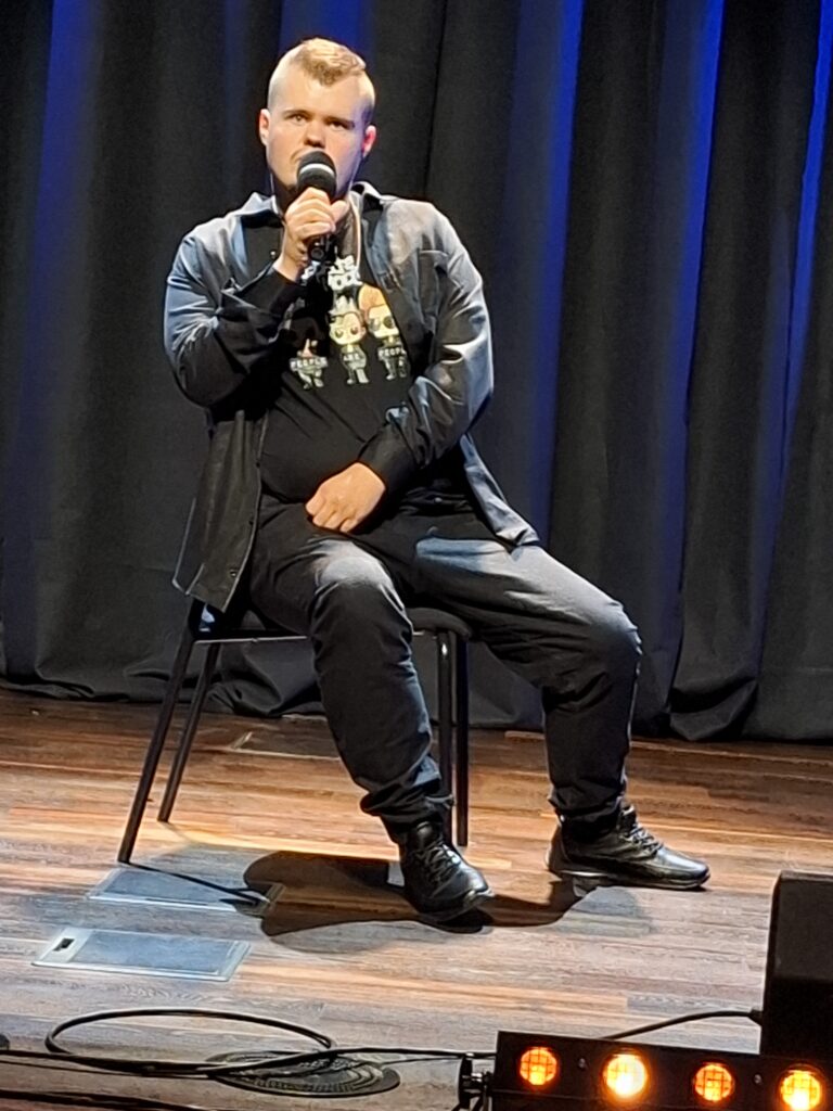 Mężczyzna siedzi na krześle na scenie i śpiewa do mikrofonu podczas festiwalu.